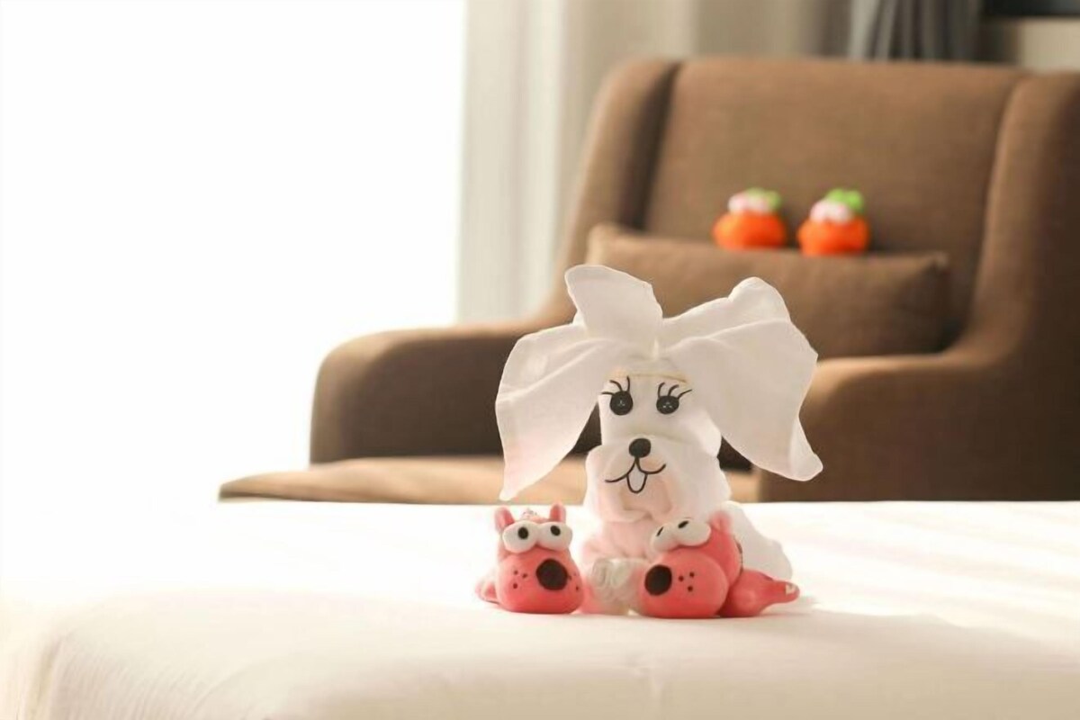 Un jouet en peluche avec un nœud, posé sur un lit dans une pièce où se trouve un canapé marron et deux petits objets rouges à l’arrière-plan.