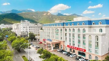 Hotel com um edifício grande, estacionamento e montanhas ao fundo.