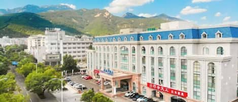 A large building with a blue roof and multiple windows, surrounded by greenery and a mountainous backdrop.