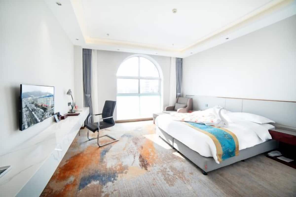 A modern hotel room with a large bed, a desk, a chair, a TV, and a large window.