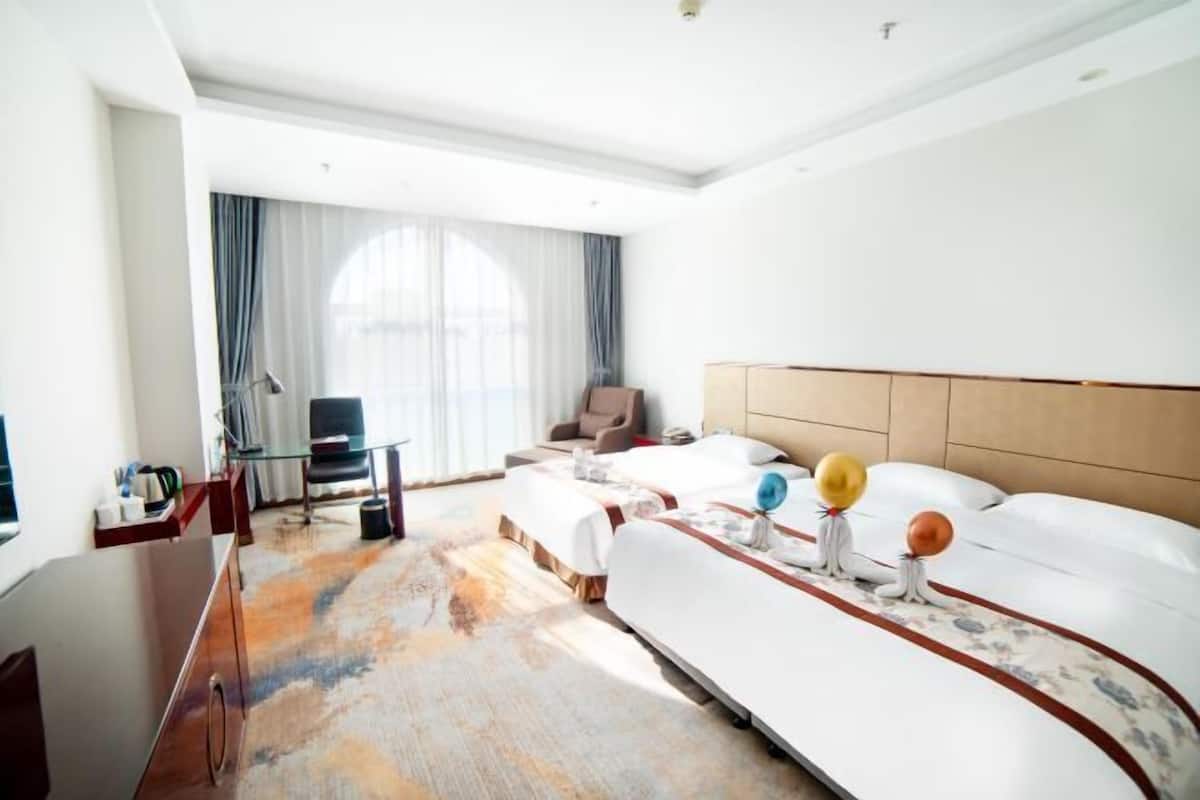 A hotel room with two beds, a desk, a chair, a large window with curtains, and a view of the outdoors.