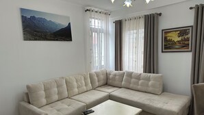 Un salon avec un canapé d’angle beige, une table basse blanche et un tapis. Il y a deux fenêtres avec des rideaux, un tableau au mur et un lustre.