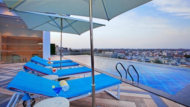 A rooftop pool area with blue lounge chairs and umbrellas, a glass balustrade, and a city view in the background. - Country Inn Premier Amritsar (Amritsar)