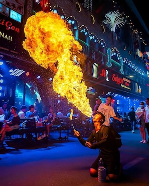 Escena callejera con un artista malabarista lanzando antorchas encendidas, rodeado de gente y letreros iluminados.