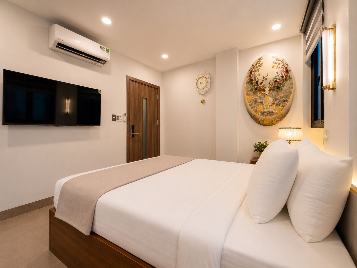 A hotel room with a bed, air conditioning unit, wall-mounted TV, wooden door, and a decorative wall piece.