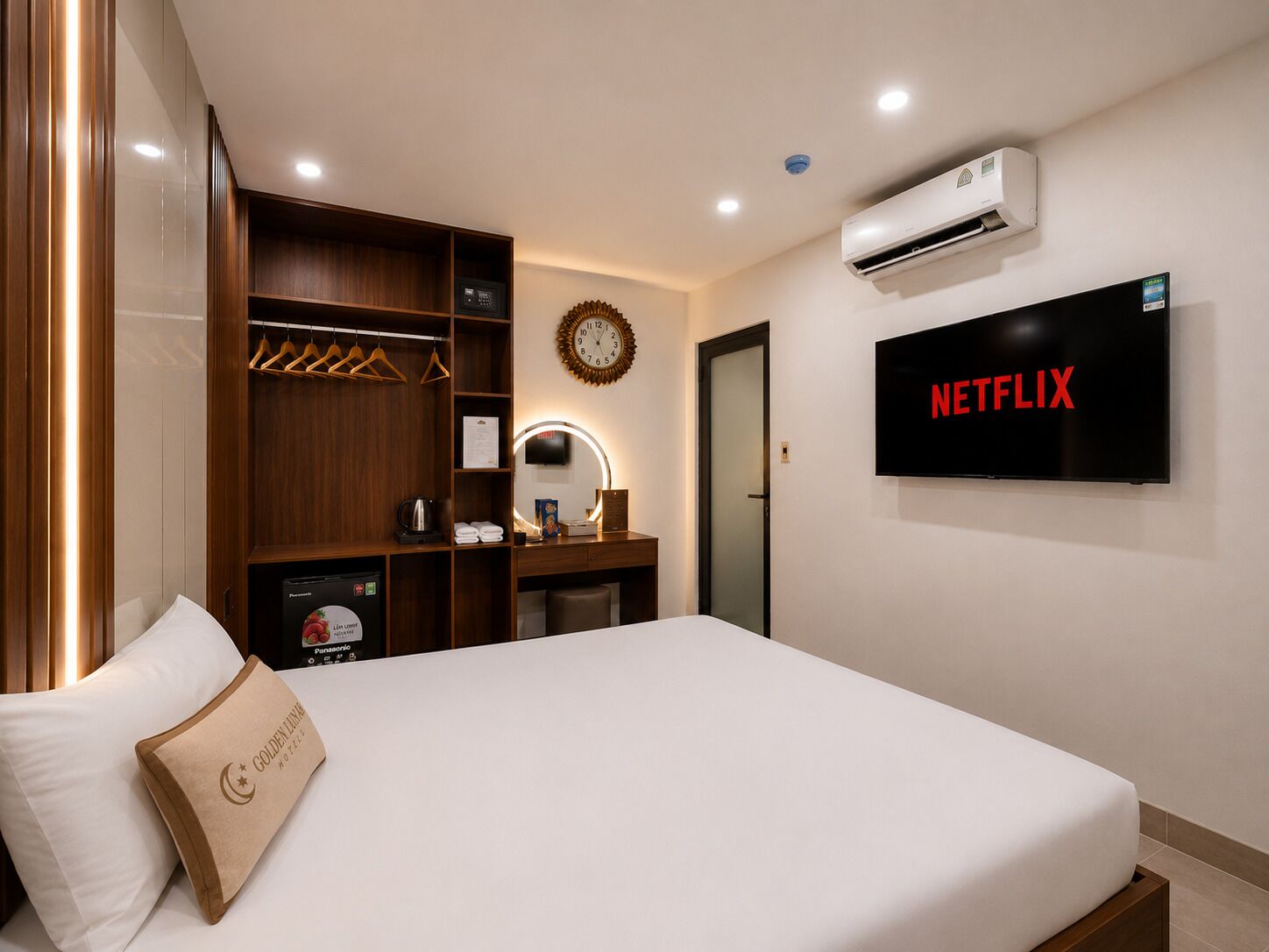 A hotel room with a bed, a wardrobe, a vanity with a mirror, a clock, an air conditioning unit, and a television displaying 'NETFLIX'.