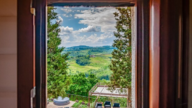 A window view of a green landscape with hills and trees, a small building with a roof, and two chairs outside.