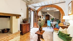 A spacious living area with a wooden bench, a sofa, and a dining table with chairs. An archway leads to a kitchen area.