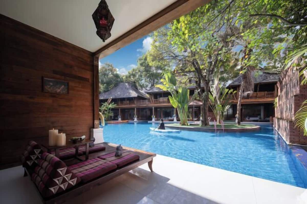 A poolside area with a wooden bench and cushions, a clear view of a swimming pool, and traditional-style buildings in the background.