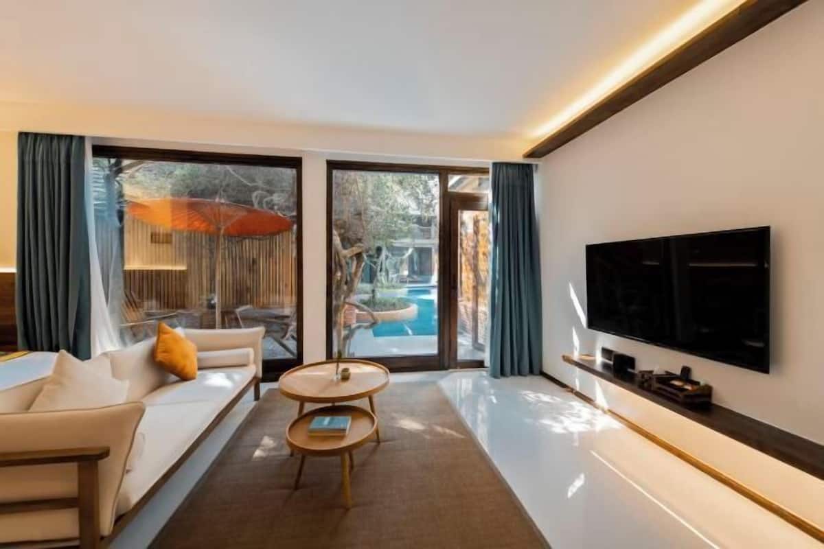A modern living room with a sofa, coffee table, and TV, large windows, and a pool view.