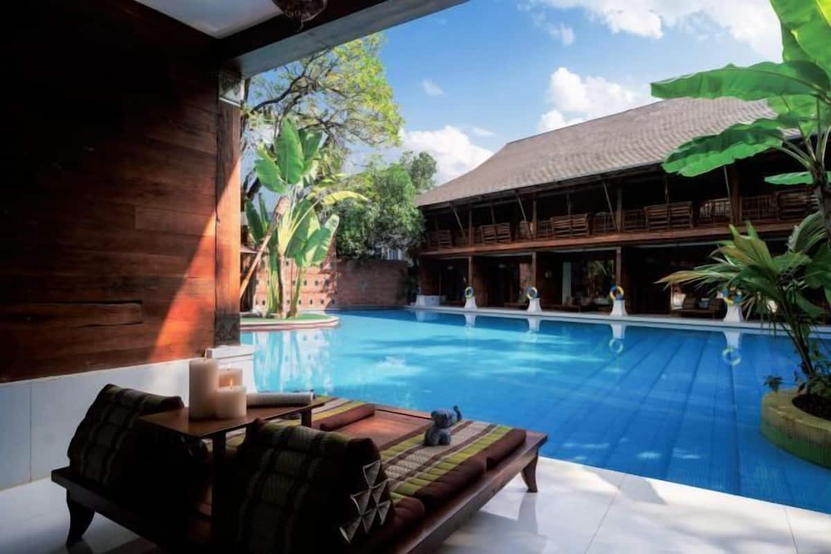 A poolside area with a wooden deck, a cushioned seating area, and a view of a building with a covered patio.