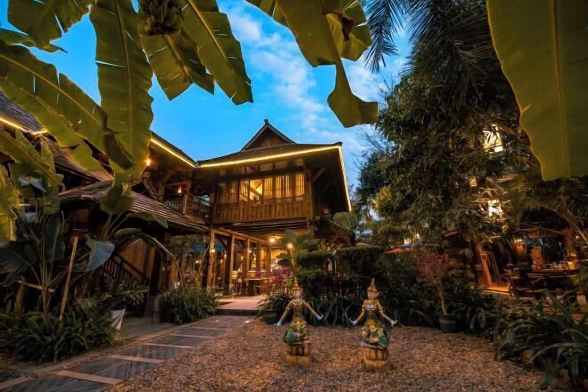 A traditional wooden building with a covered patio area, surrounded by lush tropical plants and decorative statues.