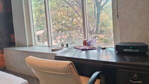 A room with a desk, a chair, a window with a view of trees, and a clock on the desk. - Stylish and Cosy Studio with huge window view, close to Moolchand Metro Station (New Delhi)