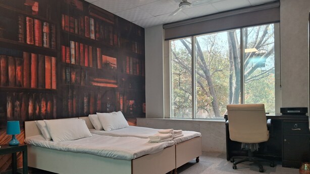 A bedroom with a large bed, a desk, and a chair. There is a window with a view of trees. - Stylish and Cosy Studio with huge window view, close to Moolchand Metro Station (New Delhi)