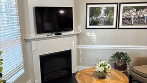 Una sala de estar con chimenea, televisor montado arriba, una mesa de centro redonda de madera con flores y un sofá.