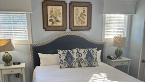 Un dormitorio ordenado con una cama grande, cabecera, dos lámparas de pared y obras de arte botánicas enmarcadas.