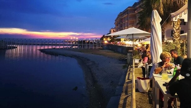 Ein Restaurant direkt am Wasser mit Außenbereich, das auf ein ruhiges Gewässer und einen Sonnenuntergang blickt.
