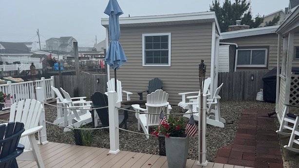 A wooden deck with white Adirondack chairs, a small potted plant with American flags, and a blue umbrella. - Lagoon Life, Beach Please! (Stafford Township)