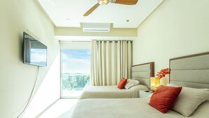 Habitación de hotel con dos camas, ventilador de techo, televisor montado en la pared y un ventanal con cortinas.