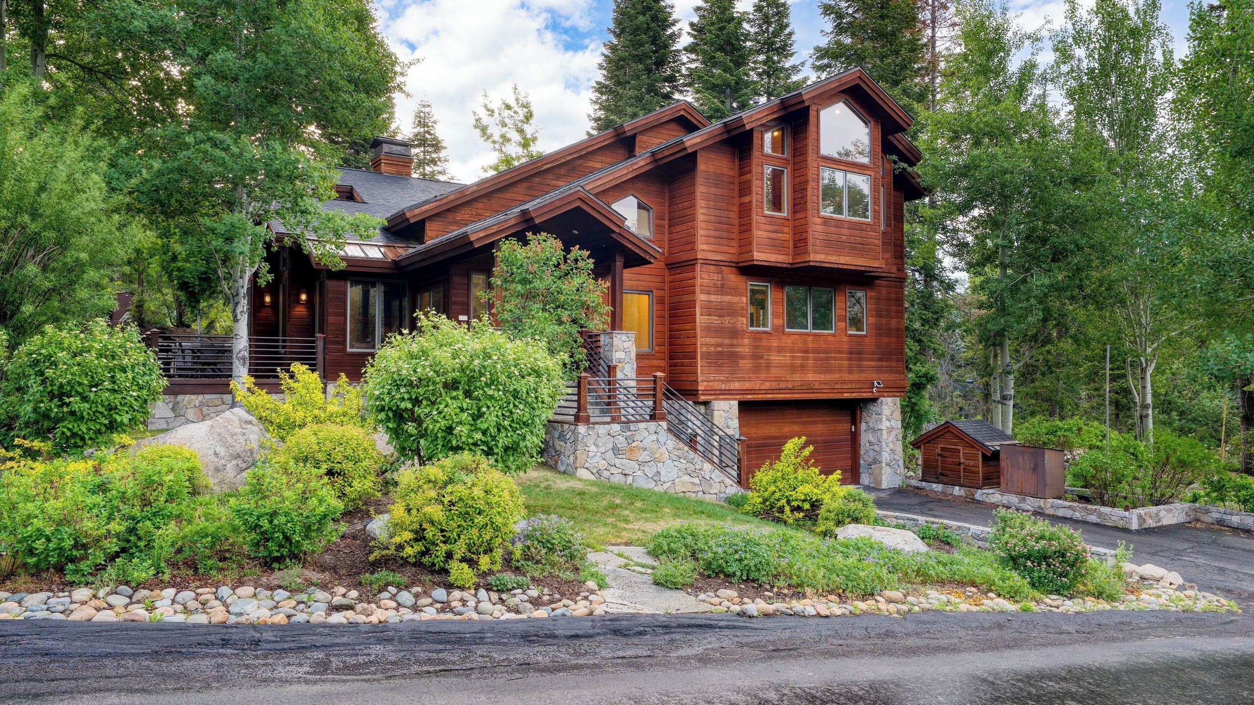 A two-story wooden cabin with a stone foundation, surrounded by lush greenery and trees.