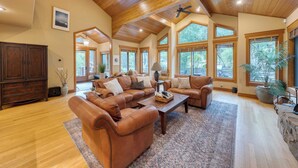 A spacious living room with leather sofas, a coffee table, and large windows offering natural light and views of the outdoors. - Cedars at Palisades by Avantstay Hot Tub, Resort Access, and Mountain Views (Olympic Valley)