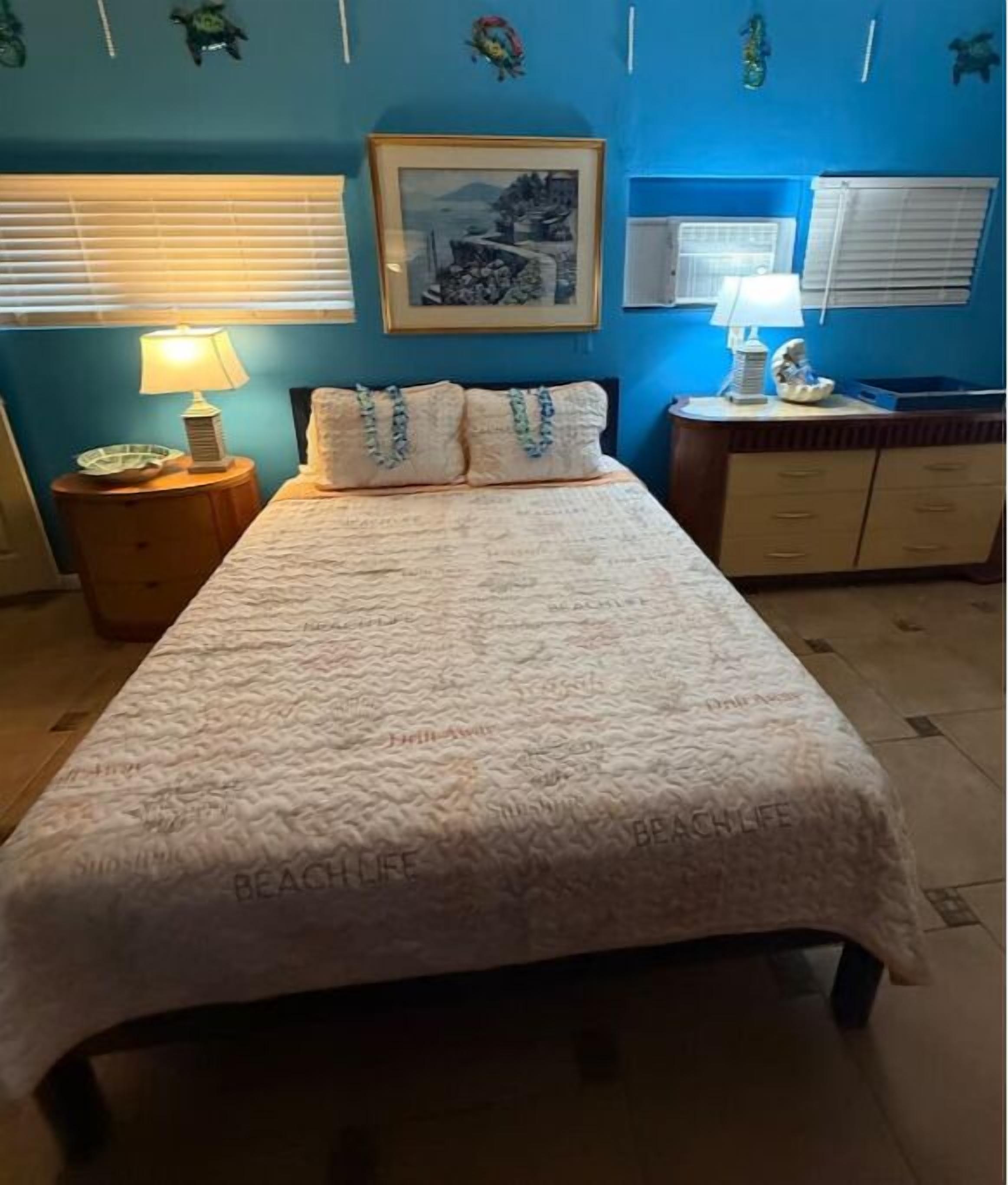 Un dormitorio con cama, mesita de noche, cómoda, un cuadro enmarcado en la pared y una ventana con persianas.