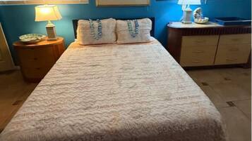 Un dormitorio con cama, mesita de noche, cómoda, un cuadro enmarcado en la pared y una ventana con persianas.
