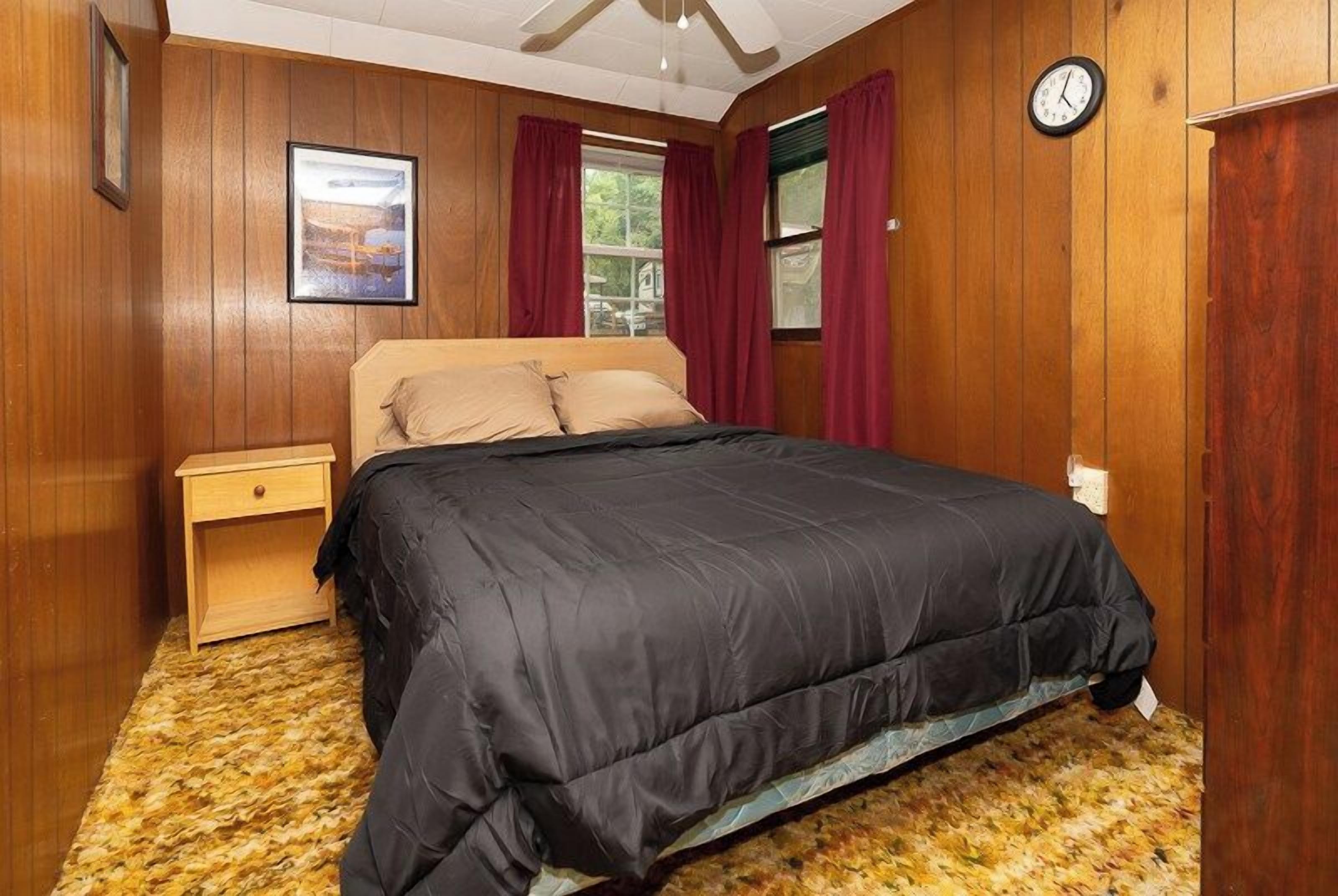 Quarto com cama, paredes de madeira, mesa de cabeceira, relógio e janela com cortinas.