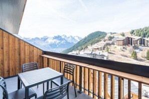 Een houten balkon met een tafel en stoelen, met uitzicht op een bergachtig landschap met gebouwen.