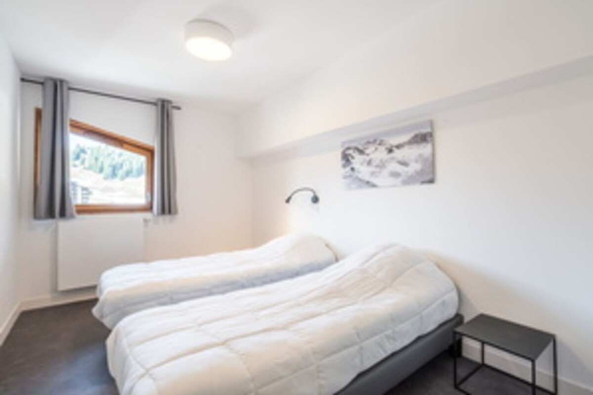 Een kamer met twee bedden, een raam met gordijnen, een wandlamp en een klein tafeltje.