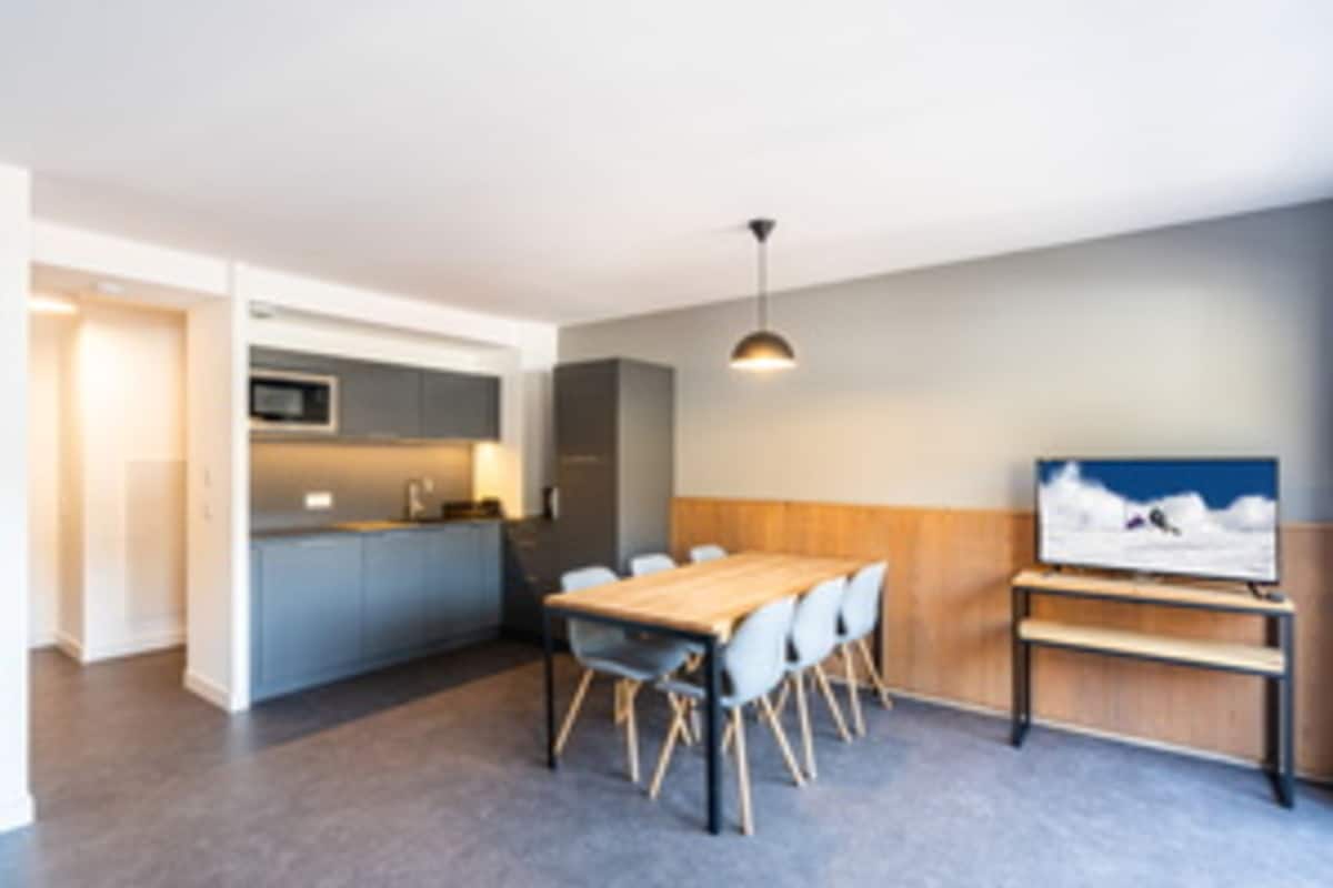 A modern kitchen with grey cabinets, a wooden dining table, and a TV on the wall.