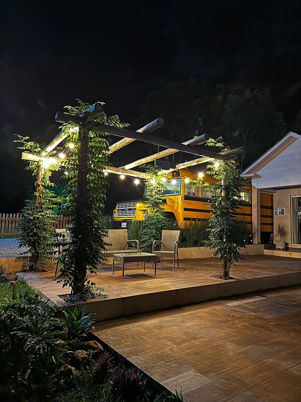 A patio area with outdoor seating, a pergola with string lights, and a yellow building in the background. - American Bus Glamping (Capinzal)