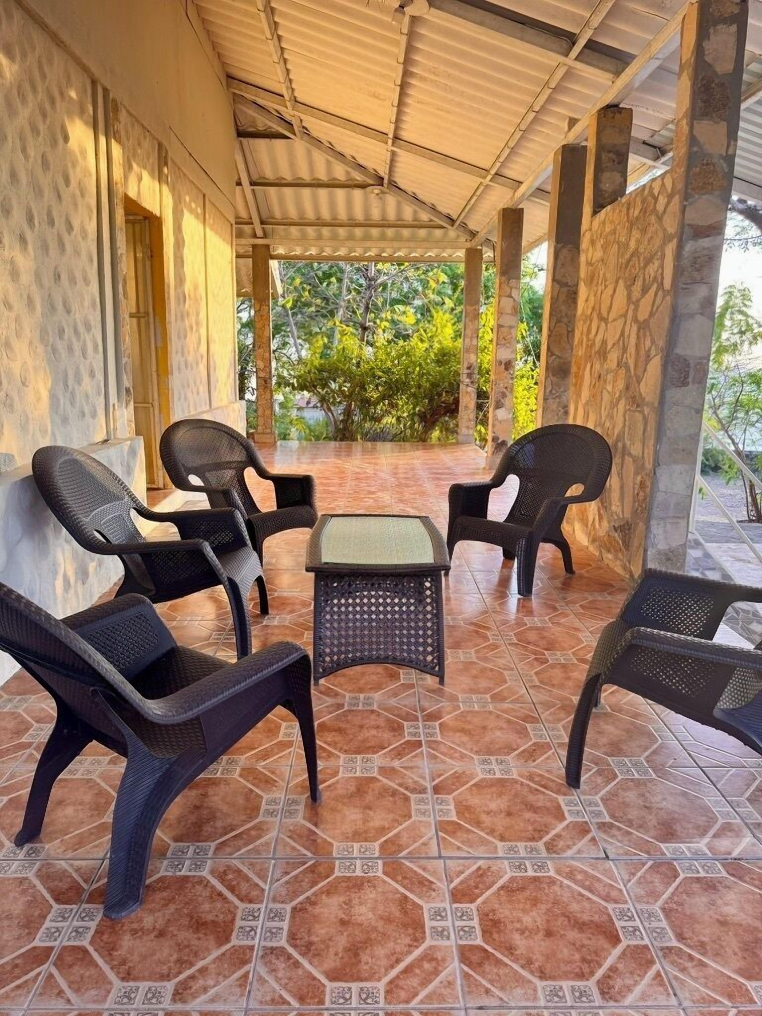 A covered outdoor seating area with wicker chairs and a small table, surrounded by stone walls and a tiled floor.