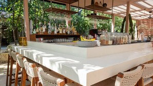 An outdoor bar area with a long white counter, wicker chairs, hanging lights, and a variety of bottles and glasses. - Resort-Style Loft in AfloraTulum (Tulum)