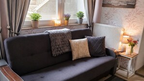 A dark-colored sofa with patterned cushions, a small side table with a lamp and flowers, and a window with curtains and potted plants.