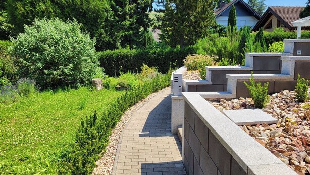 Un chemin pavé, bordé d’un côté par un mur de pierres et de l’autre par une zone herbeuse plantée d’arbres.