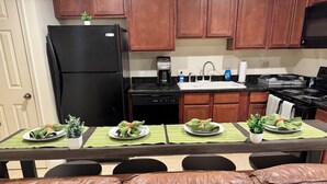 Un comedor con una mesa dispuesta para cuatro personas, un refrigerador negro, armarios de madera y una cafetera.