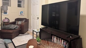 Una sala de estar con un televisor de pantalla plana grande, un sofá de cuero marrón, una mesita auxiliar pequeña y una estantería llena de DVDs.