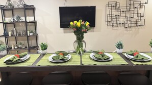 Un comedor con una mesa larga dispuesta para cuatro, un florero con flores amarillas y un televisor montado en la pared.
