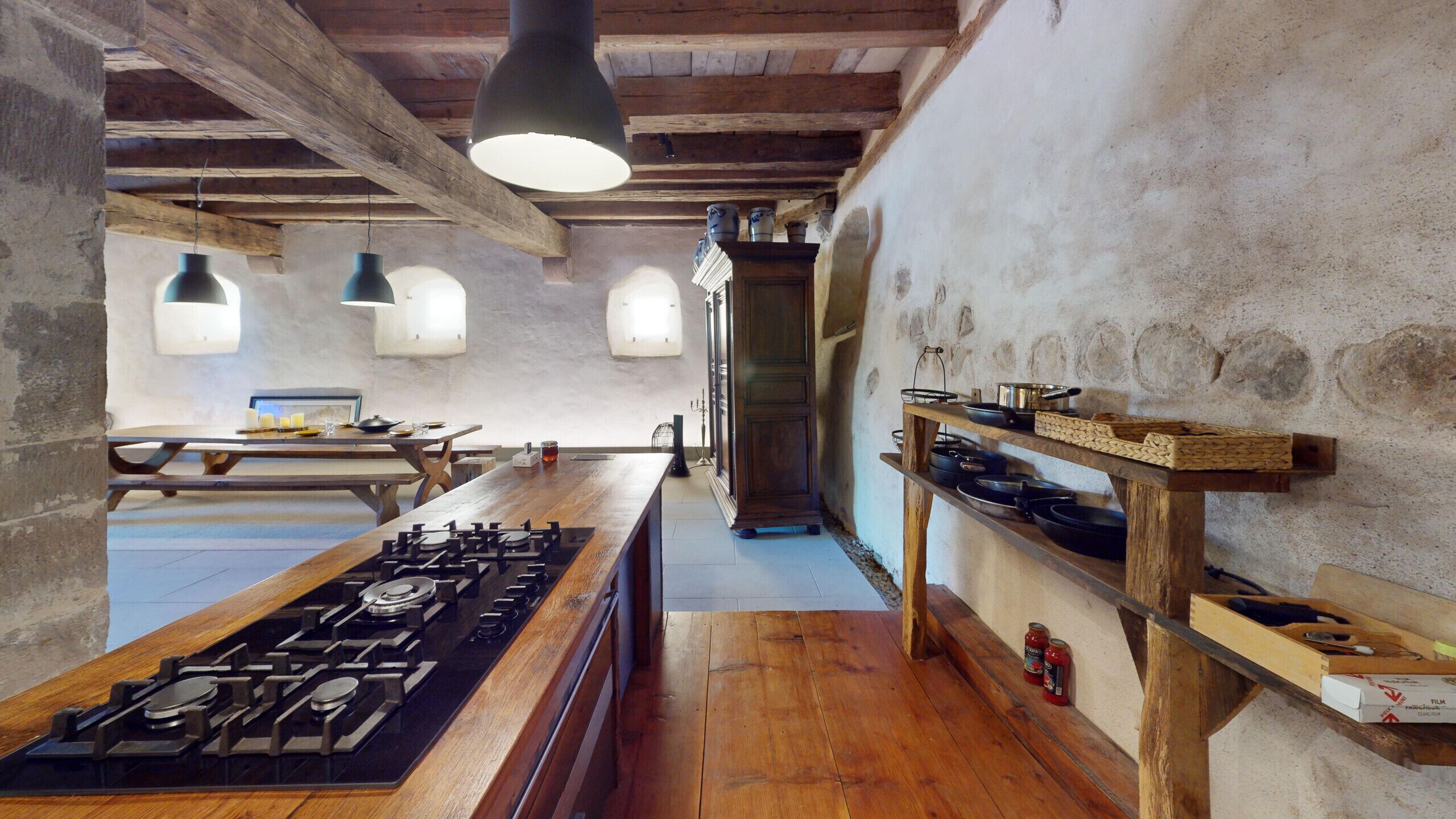 Una cocina rústica con una mesa de madera, estufa a gas, lámparas colgantes y estantes con utensilios de cocina.