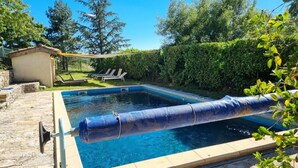 Una piscina rectangular con una cubierta azul, rodeada por un suelo de piedra y un pequeño cobertizo.