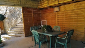 Un patio exterior con una mesa de madera y cuatro sillas, una planta en maceta y una escalera de piedra que lleva a una puerta de madera.
