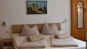 Una cama bien tendida con almohadas estampadas y toallas dobladas, un cuadro de un paisaje montañoso en la pared y una puerta de madera con un espejo.