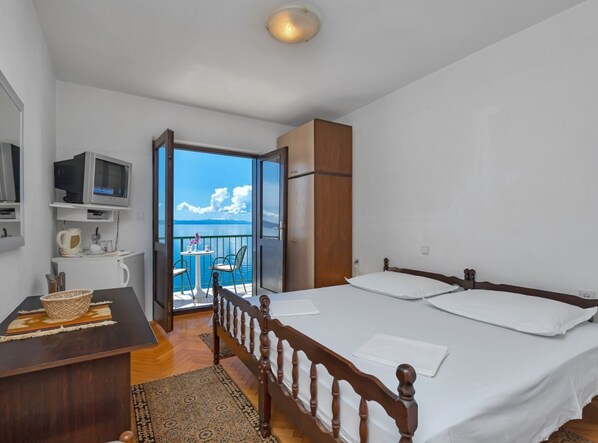 A bedroom with a wooden bed, a desk with a basket, a television, and a balcony with a view of the sea.