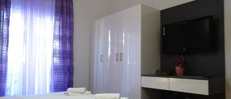 Habitación de hotel con cama, armario, escritorio con televisor de pantalla plana y ventana con cortinas moradas.