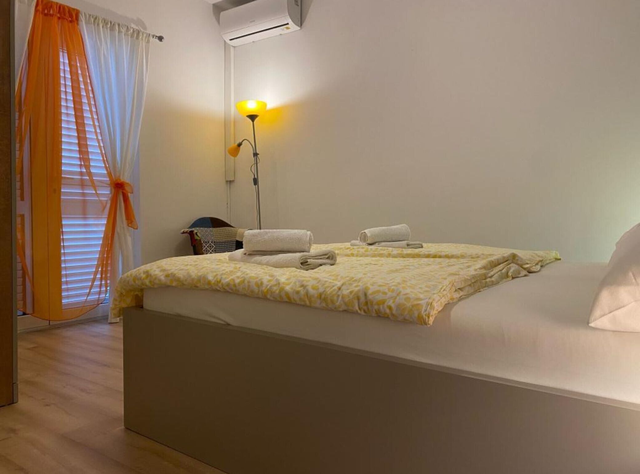 A hotel room with a bed, a bedside lamp, a window with blinds, and an air conditioning unit.
