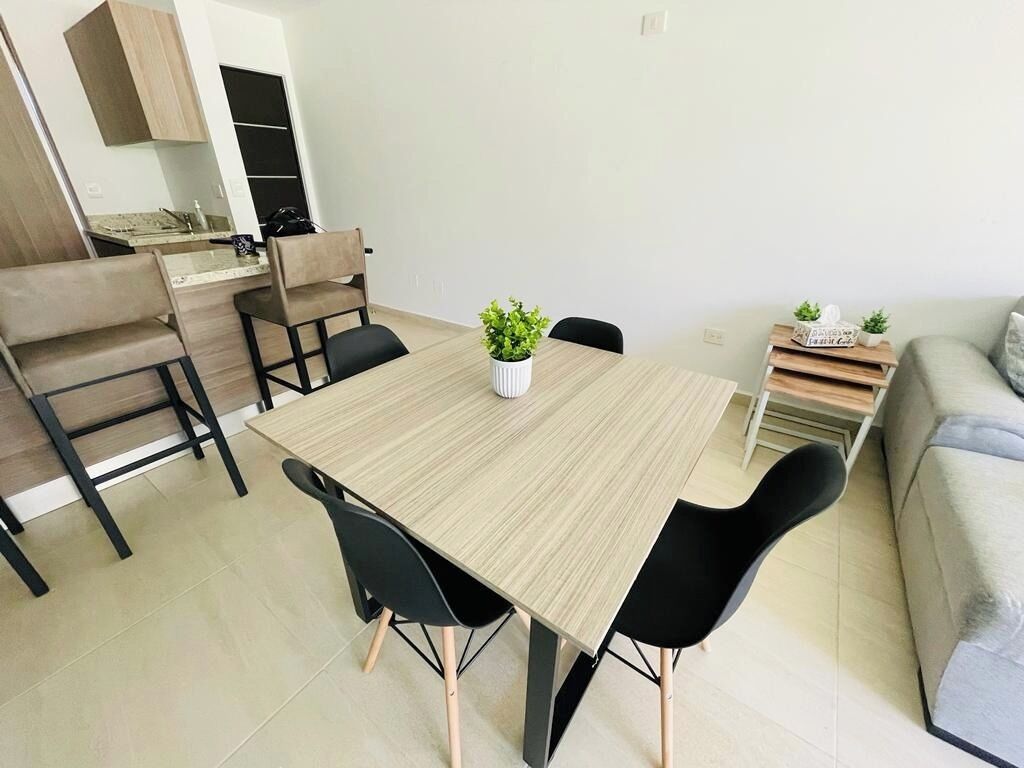 A modern dining area with a wooden table and black chairs, a small potted plant, and a kitchen area in the background.