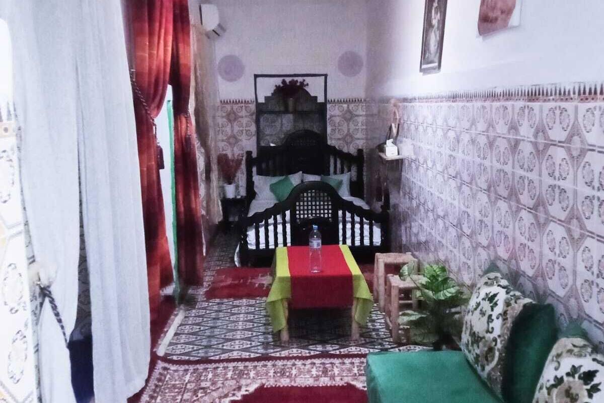 A room with a bed, a green sofa, a small table with a red cloth, patterned walls, and a patterned rug.