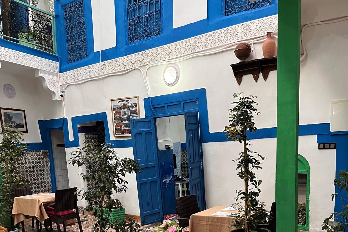 A courtyard with a blue and white building, potted plants, a table with a chair, and a patterned rug.