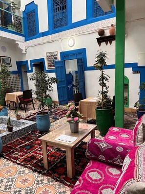 A courtyard with a blue and white building, potted plants, a table with a chair, and a patterned rug. - Riad dar Medina Land (Marrakech)